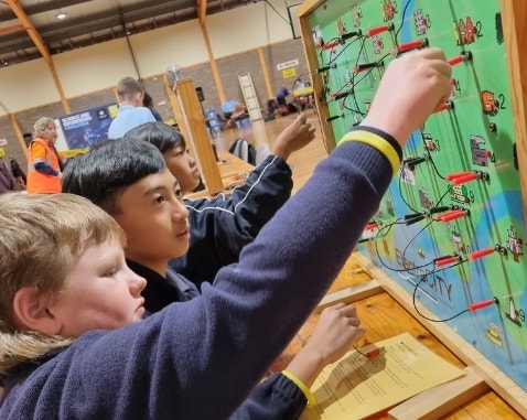 Photo of three students looking at a display circuit board, reaching up to touch it